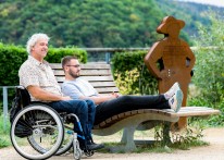 Foto: Zwei M&auml;nner sitzen im Freien, einer auf einer Bank, der andere im Rollstuhl